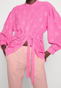 Camicia rosa testurizzata con vita annodata e chiusura a bottoni, dotata di maniche lunghe e vestibilità rilassata; abbinata a pantaloni rosa chiaro.