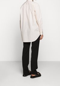 Beige raitoja omaava napillinen paita, jossa on kaareva helma, pitkät hihat ja yhden nappulan hihansuut, yhdistettynä mustiin housuihin ja remmikenkiin.