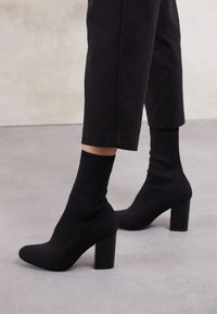 Bottes à talon en chaussettes noires en tissu extensible, dotées d'un talon bloc haut et d'un poignet côtelé, rehaussées d'une finition lisse et élégante.