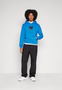 Blauwe hoodie met een voorzak, capuchon met trekkoord en een zwart logo-embleem. Gecombineerd met zwarte broek en witte sneakers.