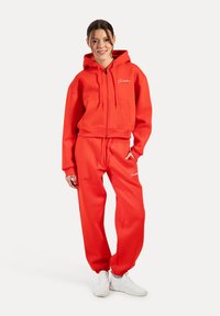 Červená mikina na zip s předními kapsami a odpovídající tepláky, které mají elastické manžety a malou logovou aplikaci na nohavicích. Bílé tenisky.