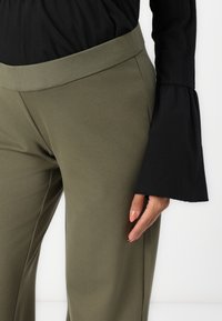 Pantaloni verde oliva con una texture liscia e una ampia vita elastica, abbinati a un top nero a maniche lunghe con polsini a campana.