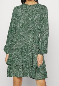 Vihreä kuosiset mekko, jossa on kukkakuviot, pitkät hihat ja kerroksellinen röyhelöhelma. Pehmeä kangas, jossa on hieman läpikuultava rakenne.