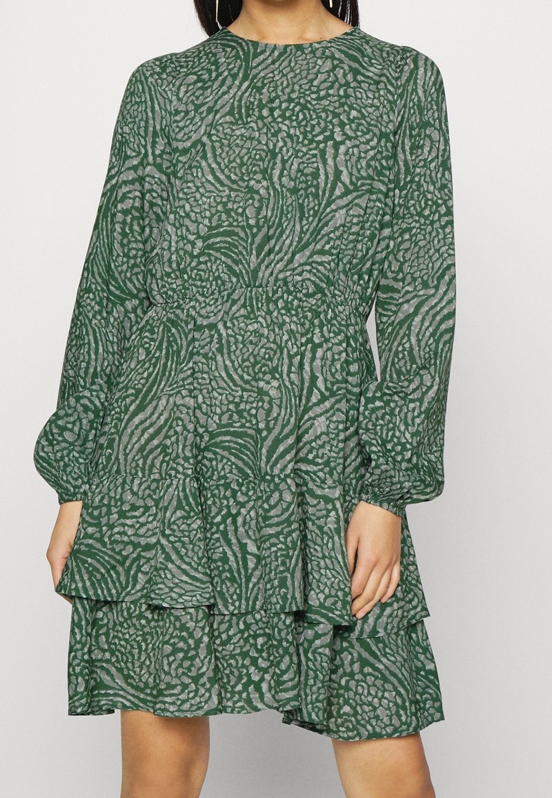 Vihreä kuosiset mekko, jossa on kukkakuviot, pitkät hihat ja kerroksellinen röyhelöhelma. Pehmeä kangas, jossa on hieman läpikuultava rakenne.