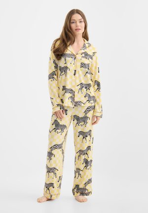 Vrouw die blootsvoets staat en gele geruite pyjama draagt bedrukt met zwart-witte zebra's tegen een effen witte achtergrond.