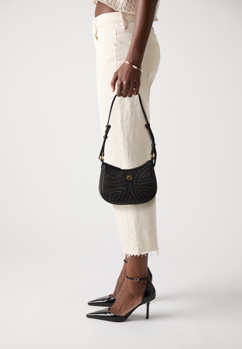 Schwarze Lederhandtasche mit strukturiertem Blumenmuster, kurzer Tragegurt, goldene Hardware; kombiniert mit weißen Jeans und schwarzen Lackleder-High Heels mit Knöchelriemen.