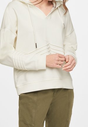 Femme portant un sweat à capuche écru avec un col en V et des détails en chevron texturés, associé à un pantalon vert olive, mains jointes près de la taille.
