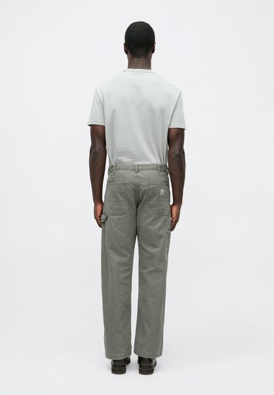 Ανοιχτό γκρι μπλουζάκι, χαλαρά χακί παντελόνια cargo με τσέπες και μαύρα παπούτσια, από πίσω, σε απλό φόντο.