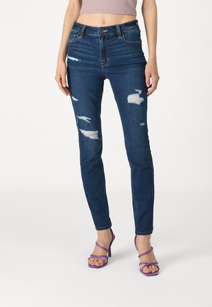 Hochgeschnittene blaue Jeans aus Denim mit verwaschenen Details und sichtbaren Nähten. Kombiniert mit lila Riemchen-Pumps.