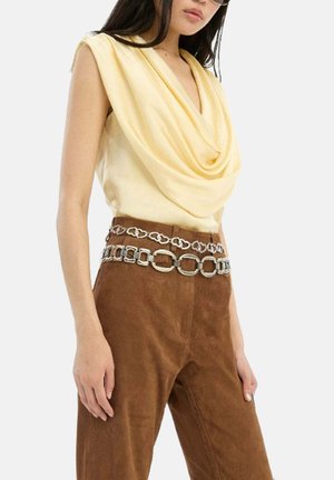 Femme portant un haut jaune sans manches drapé, un pantalon en daim marron et deux ceintures chaîne argentées à gros maillons.