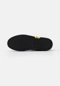 Black rubber shoe sole with a textured surface, featuring small triangular patterns, and a yellow accent with a visible brand name.