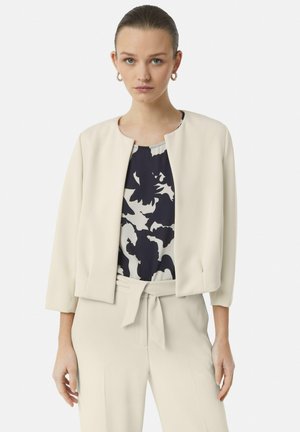 Crème-kleurige jas met een ronde hals en driekwartmouwen, gedragen over een marineblauw bloemetjestop, gecombineerd met bijpassende beige broek.