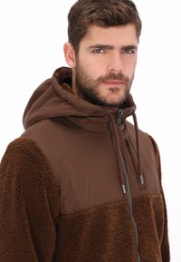 Sudadera con cremallera marrón que presenta una capa exterior suave y texturizada y un panel superior liso. Incluye capucha y cordones ajustables para un mejor ajuste.