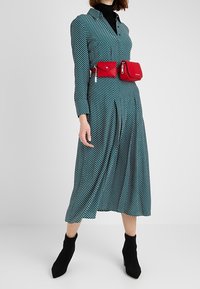 Femme portant une robe midi à pois vert sarcelle, des bottines noires, un col roulé noir et un sac ceinture rouge avec deux pochettes autour de la taille.