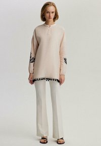 Tunic rosa chiaro a maniche lunghe con ricami neri di foglie sulle maniche e bordatura con frange nere sull'orlo, abbinata a pantaloni beige a gamba larga.