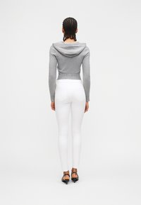 Γυναίκα με πλεξού δες στέκεται με την πλάτη, φορώντας εφαρμοστό γκρι πουλόβερ με κουκούλα, λευκά στενά τζιν και μαύρα παπούτσια με χαμηλό τακούνι.