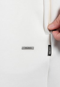 Main tenant une fermeture éclair sur un tissu blanc à proximité d'étiquettes en métal et en tissu portant le label "SMILODOX" sur un vêtement.