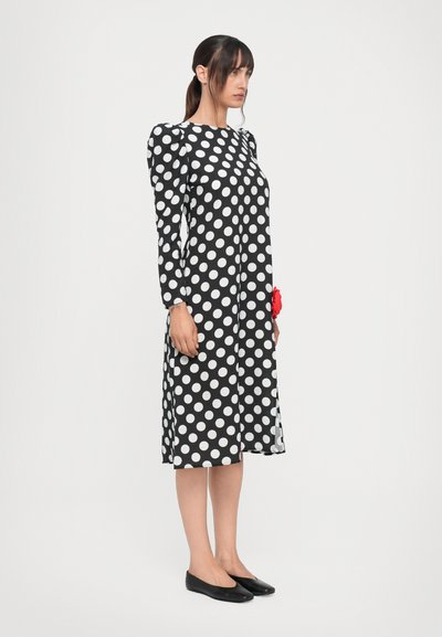 Musta-valkoinen pilkullinen mekko pitkillä hihoilla, suora leikkaus ja kerätty olkahihnasuunnittelu, yhdistettynä mustiin ballerina-kengiin. Punainen kukkaaksentti.