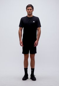 Άνδρας που στέκεται με το πρόσωπο προς τα εμπρός, φοράει μαύρο αθλητικό μπλουζάκι Adidas, σορτς, κάλτσες και μαύρα αθλητικά παπούτσια, μπροστά σε απλό λευκό φόντο.