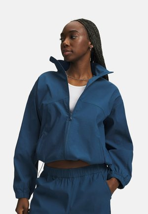 Vrouw met gevlochten haar die een blauwe jas met ritssluiting draagt over een wit topje, gecombineerd met bijpassende blauwe broek met elastische tailleband, handen in de zakken.