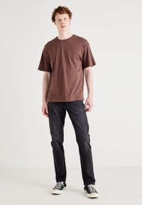 Brown cotton T-shirt with a crew neck, relaxed fit. Paired with dark denim jeans and black and white canvas sneakers.