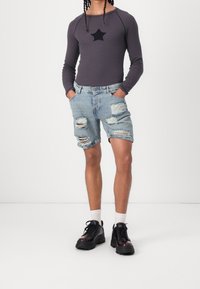 Grå långärmad skjorta med en svart stjärndetalj, i kombination med slitna blå denimshorts. Svarta klumpiga skor och vita strumpor fullända looken.