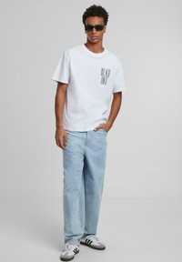 T-shirt en coton blanc avec le texte noir "REACH HIGH" sur le devant, associé à un jean bleu clair à coupe décontractée et des baskets grises à rayures.