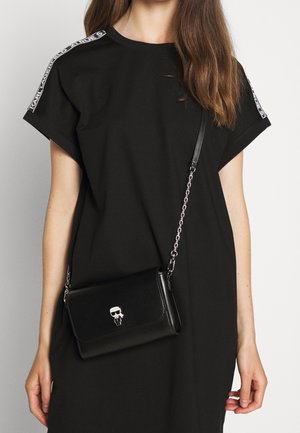 Black oversized t-shirt with short sleeves, featuring white logo detailing on the shoulders, paired with a small black crossbody bag with a skull clasp.