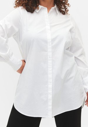 Femme aux cheveux bouclés portant une chemise blanche à manches longues et un pantalon noir, mains sur les hanches sur un fond uni.
