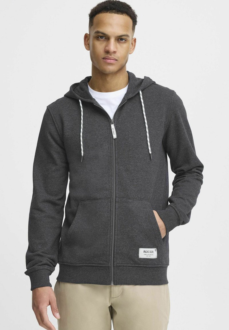 Graue Zip-Hoodie aus weichem Material, mit Kapuze, weißen Kordeln und zwei vorderen Taschen. Etikett in der unteren rechten Ecke.
