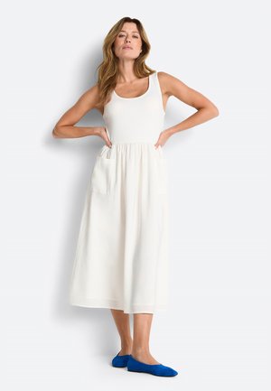 Kobieta stoi z rękami na biodrach, ubrana w białą sukienkę midi bez rękawów z przednimi kieszeniami oraz niebieskie płaskie buty na białym tle.