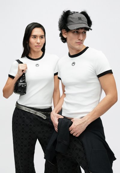 Bílé trička s černým lemováním a logem, doplněná černými kalhotami se vzorem a černou čepicí s kožešinovým zdobením. Doplňky zahrnují malou černou kabelku.