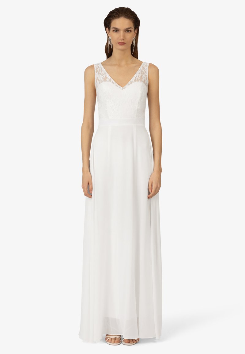 Weißes Kleid mit einem Spitzenoberteil und V-Ausschnitt, fließendem Rock und transparenten Trägern. Akzentuiert mit schlichter, eleganter Fußbekleidung.