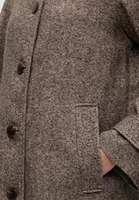Manteau en laine marron chevron avec boutons foncés, montrant un détail de face et une main partiellement dans une poche latérale.