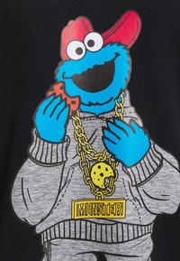 Personnage bleu portant une casquette rouge et un sweat à capuche gris, tenant un biscuit, avec une grosse chaîne en or ornée d'un pendentif biscuit et d'un pendentif "MONSTER".