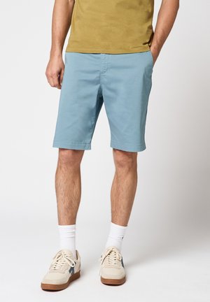 Homme portant un short bleu clair mi-cuisse, des chaussettes blanches montantes, des baskets beige avec des accents foncés, et un t-shirt jaune moutarde, debout les mains dans les poches.