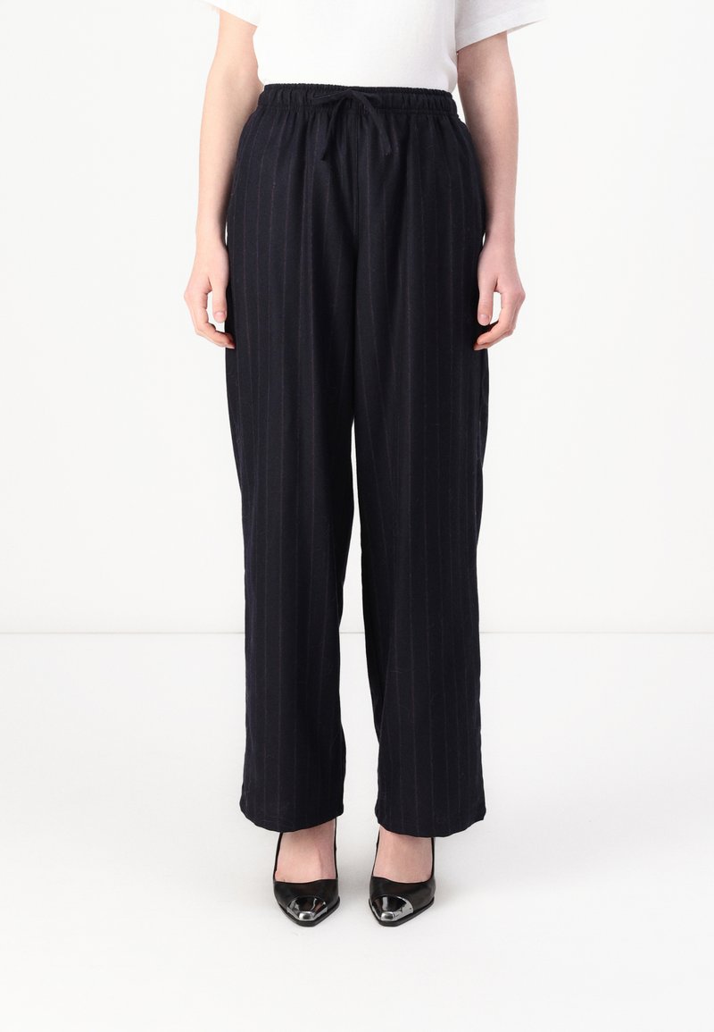Navy pinstriped trousers with an elastic waistband, relaxed fit, and side pockets, paired with black pointed-toe shoes.