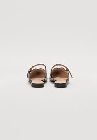 Chaussures à enfiler en cuir verni noir avec un petit talon, présentant un bord festonné et un petit détail de boucle sur la sangle. Doublure intérieure lisse.