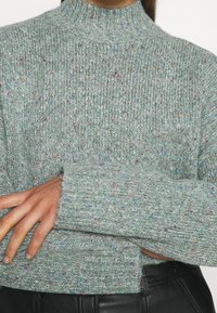 Suéter de punto en verde claro con motas multicolor, cuello alto y textura acanalada, con mangas largas y ajuste holgado.