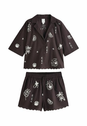 Osoba s rukou v vrecku, ktorá nosí tmavé šortky s bielymi vyšívanými vzormi, vrátane usmievavej tváre, plátku citrusov a kvetov.