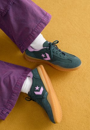 Benen in paarse broeken en witte sokken met zwarte en roze sneakers op een mosterdgeel tapijt.