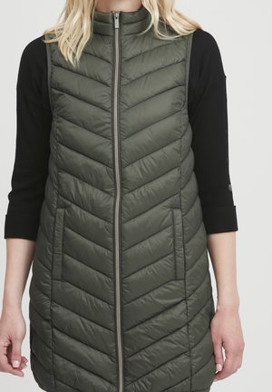 Femme portant une veste matelassée sans manches vert olive avec une fermeture éclair devant, sur un haut noir à manches trois-quarts.