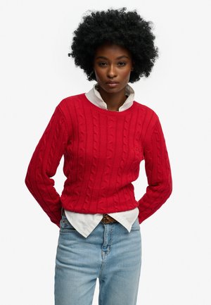 Suéter rojo de punto trenzado con cuello acanalado, usado sobre una camisa blanca con cuello. Combinado con jeans azul claro. Diseño simple y texturizado.