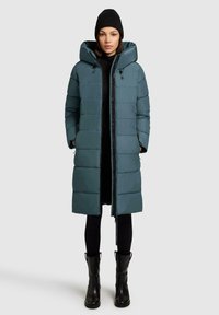 Lang pufferjakke i teal med lynlåslukning, stor hætte og quiltet design. Kombination med sorte leggings og knæhøje sorte støvler.