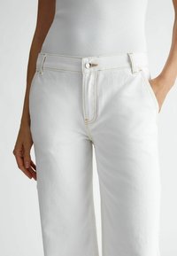 Białe dżinsy o prostym kroju, z kontrastującym żółtym przeszyciem, metalowym zapięciem na guzik oraz kieszeniami z przodu.