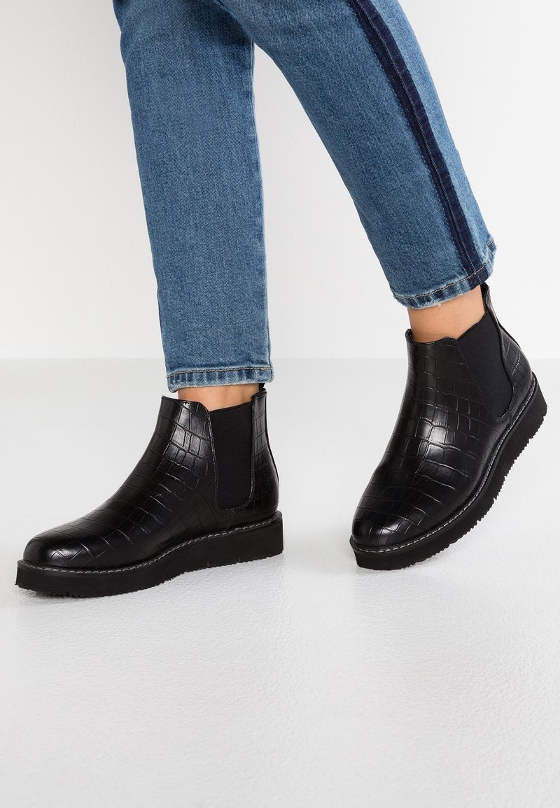 Jambes portant un jean bleu avec des bandes latérales foncées et des bottines noires à motif crocodile avec des panneaux latéraux élastiques sur un sol blanc.