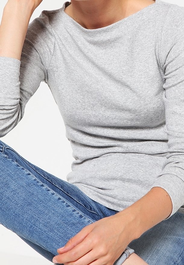 Person som bär en ljusgrå långärmad tröja och blå jeans, sitter med benen i kors mot en vit bakgrund.