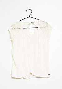 Blouse blanche à col en V, manches courtes en dentelle, fronces subtiles à l'avant, et patte de boutonnage, suspendue à un cintre noir.