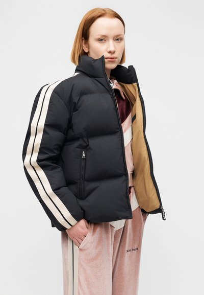 Μαύρο μπουφάν puffer με κρεμ ρίγες στα μανίκια, κλείσιμο με φερμουάρ και μπεζ φόδρα. Φορεμένο πάνω από φόρμα βελουτέ με τσέπες.