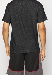 Hombre con una camiseta de manga corta gris oscuro y pantalones cortos deportivos de malla negros y grises con ribetes rojos, visto desde la espalda.